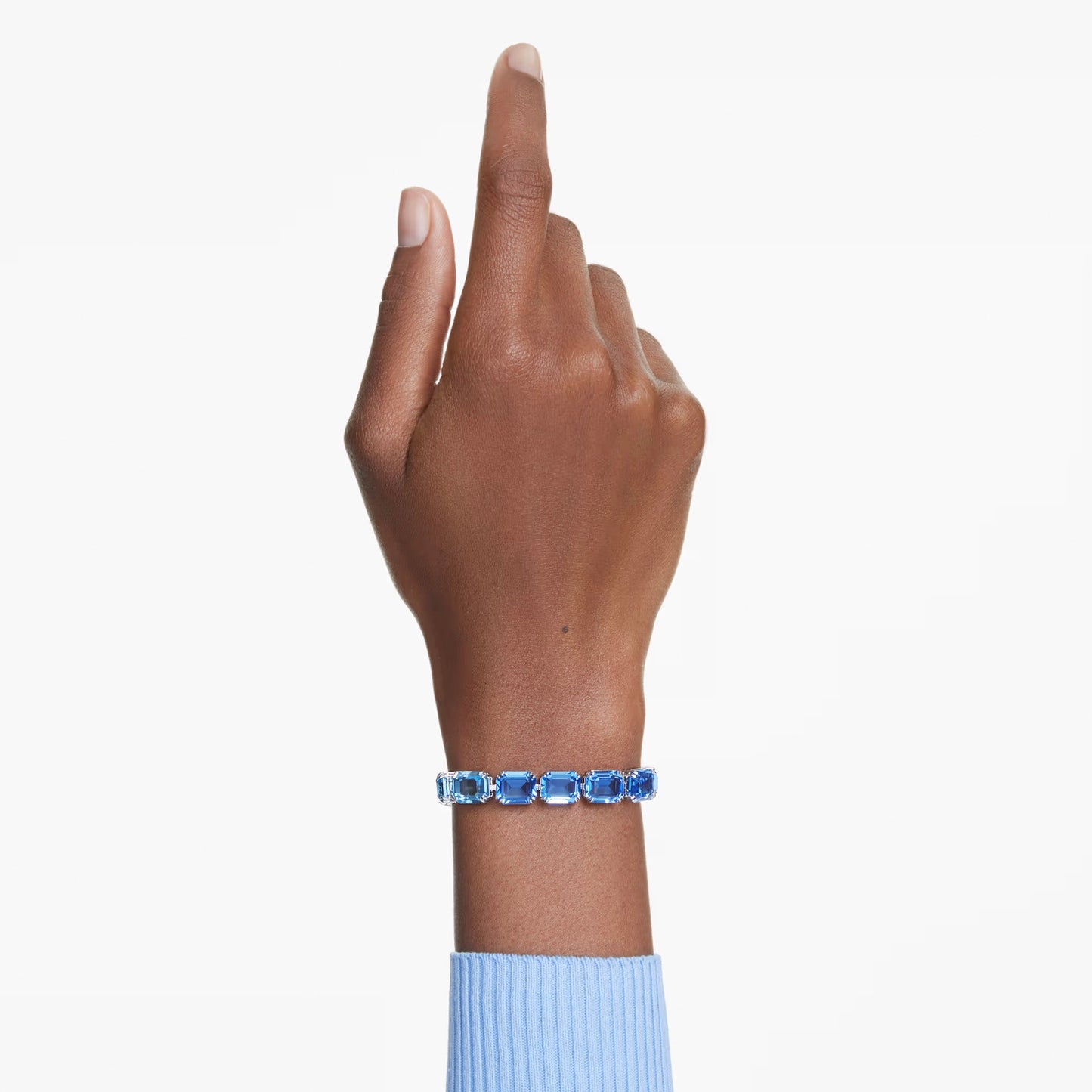 A hand pointing upward, adorned with a Swarovski Millenia Tennis Bracelet featuring Swarovski ReCreated crystals, complements a blue sweater against a light background.