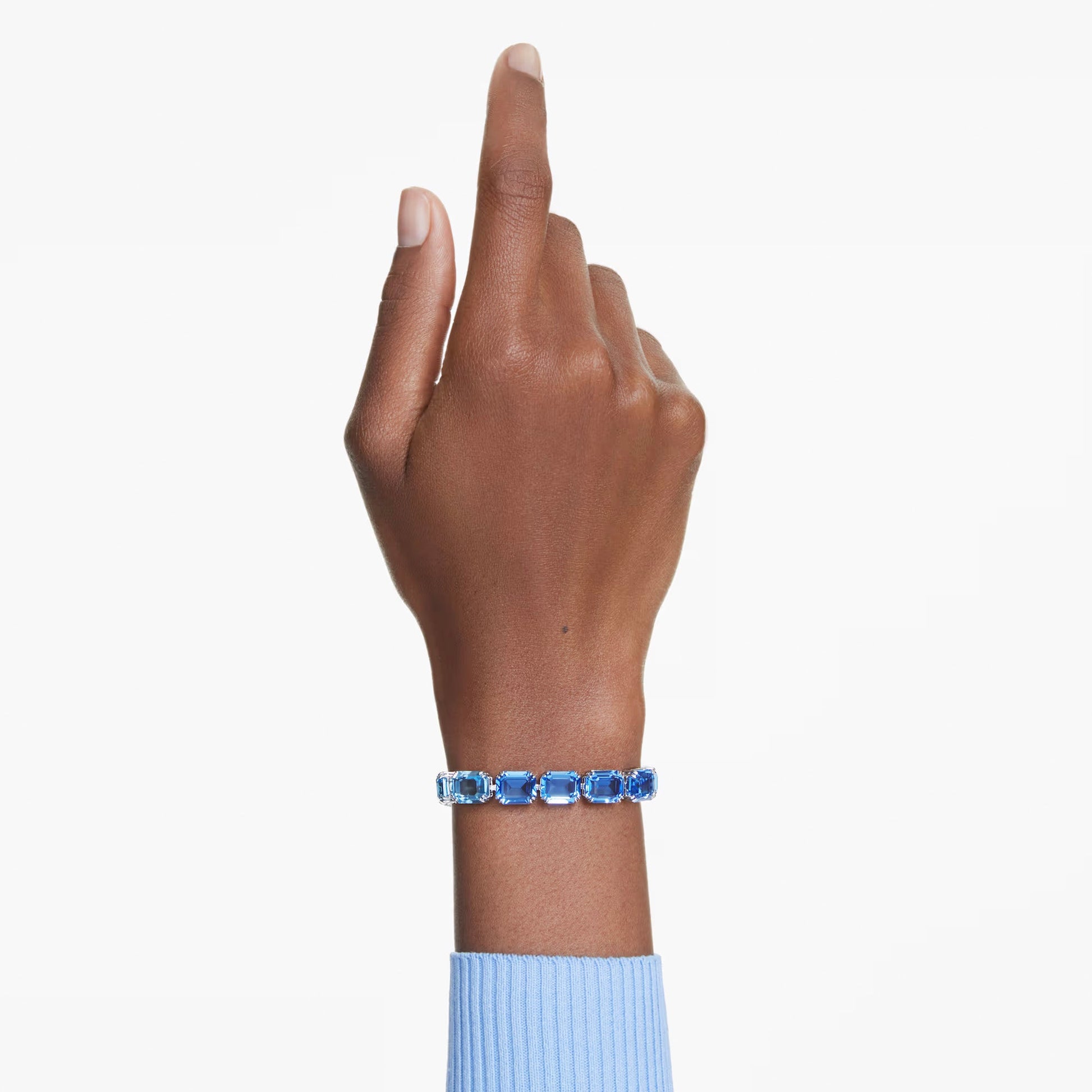 A hand pointing upward, adorned with a Swarovski Millenia Tennis Bracelet featuring Swarovski ReCreated crystals, complements a blue sweater against a light background.