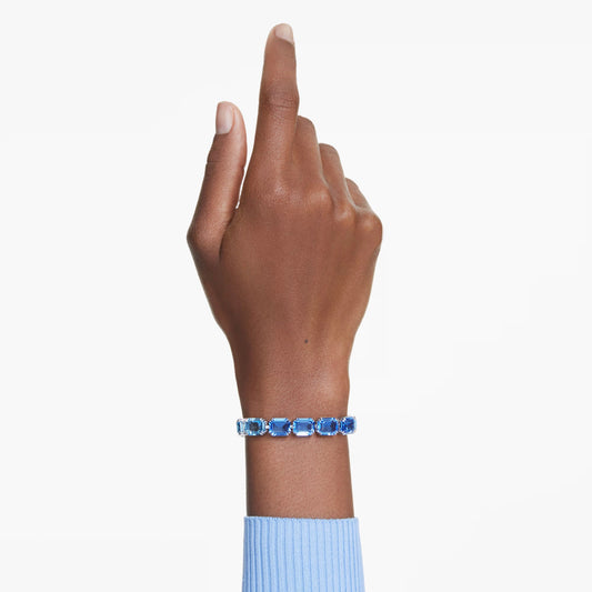 A hand pointing upward, adorned with a Swarovski Millenia Tennis Bracelet featuring Swarovski ReCreated crystals, complements a blue sweater against a light background.