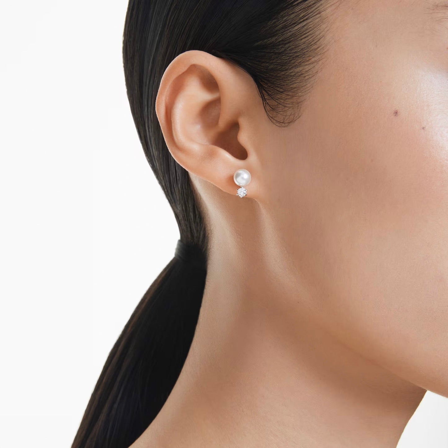 The image features a close-up of a person's ear wearing Swarovski Matrix Stud Earrings. Their hair is styled in a sleek ponytail, highlighting smooth and glowing skin, all set against a simple white background.