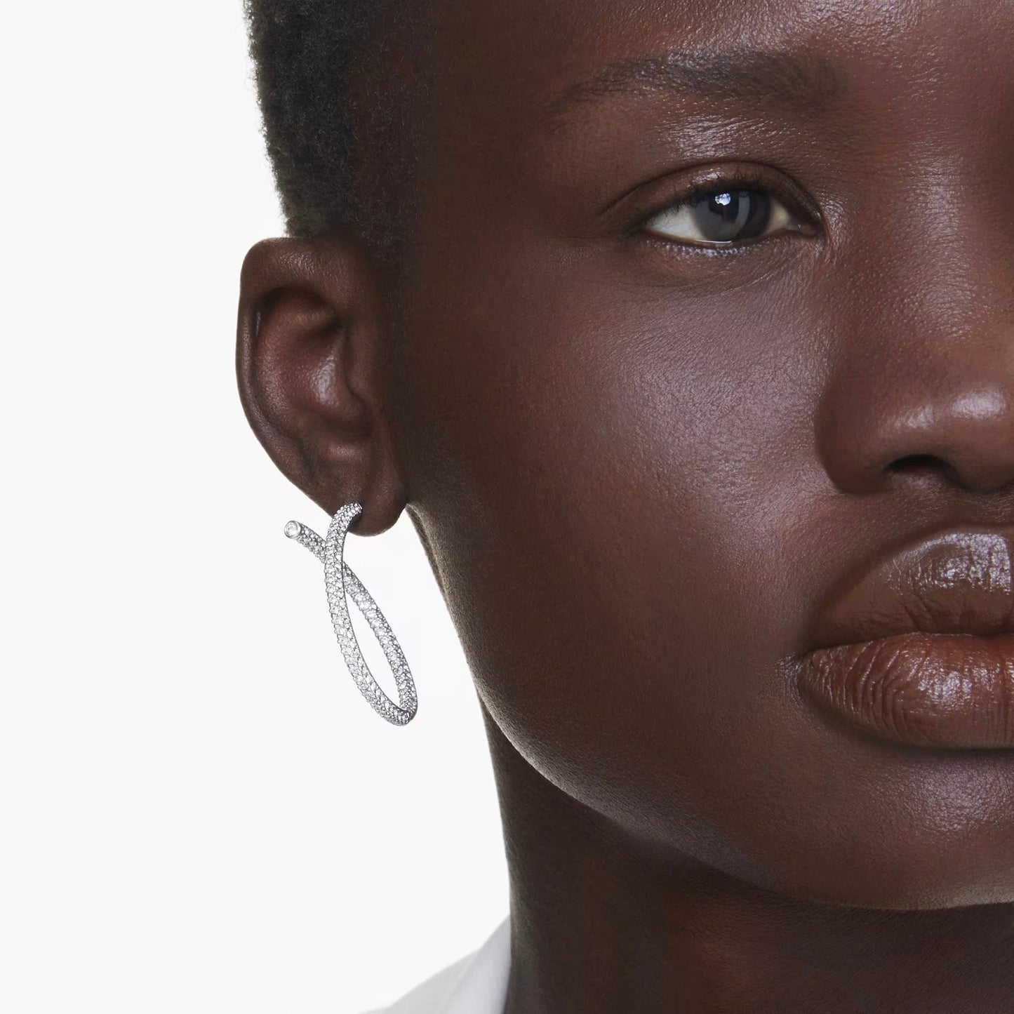 A close-up of someone wearing the eye-catching Swarovski Dextera Hoop Earrings, showcasing rhodium-plated hoops with a textured design. Their short hair and smooth, dark skin contrast beautifully against the plain white background.