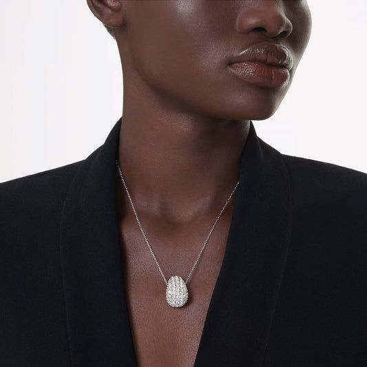 A person wearing a black jacket showcases the Swarovski Sublima Necklace Pendant, featuring dazzling clear crystals on a teardrop-shaped, silver chain. The plain, light-colored background accentuates the elegance of this stunning jewelry piece.