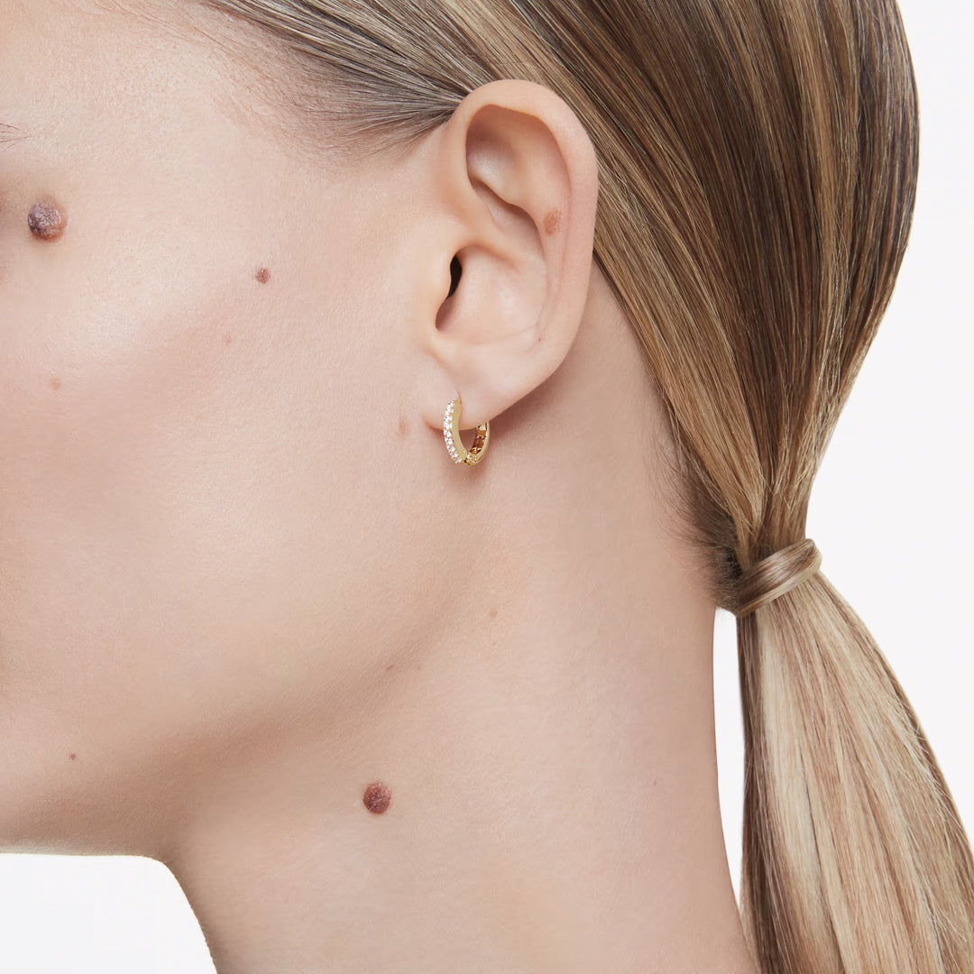 Close-up of a person with light hair tied in a low ponytail, wearing the elegant Swarovski Matrix Hoop Earrings with gold-tone plating. A few prominent moles are visible on their skin against the white background.