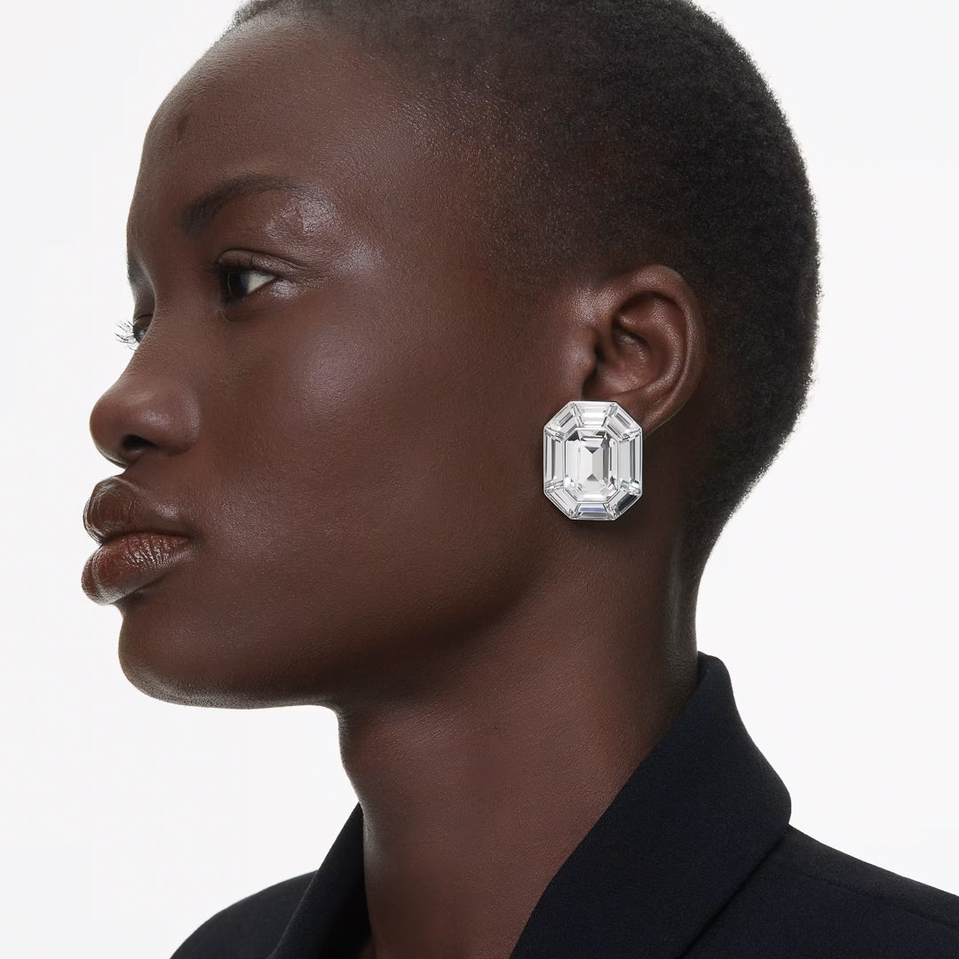 A person with short hair is shown in profile, wearing a large, octagon-cut clear crystal earring from the Swarovski Lucent Clip Earrings collection by Swarovski. They are dressed in a dark blazer against a plain white background.