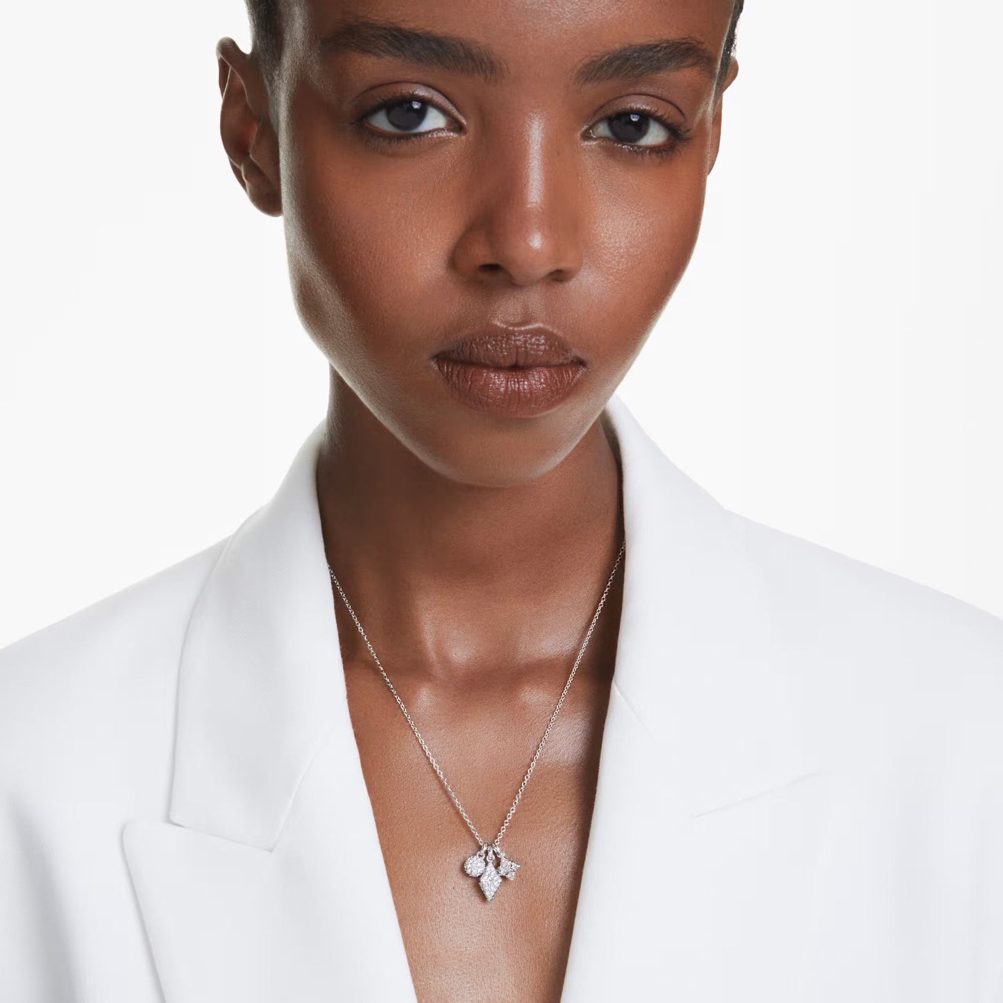 A person with short hair is wearing a white blazer and the Swarovski Dextera Necklace Pendant, which features a geometric design adorned with pavé crystals. The background is plain white.