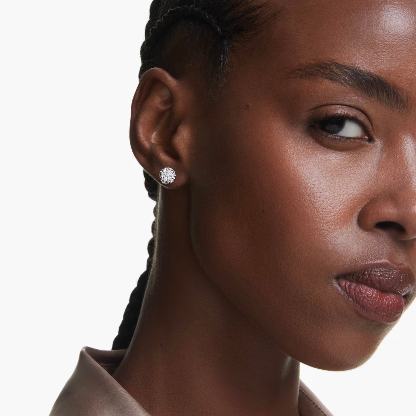 A close-up of a woman's face highlights her right side. She has braided hair and wears Swarovski Dextera Stud Earrings, beautifully accentuated with crystals. Her expression is calm and confident, set against a plain white background.