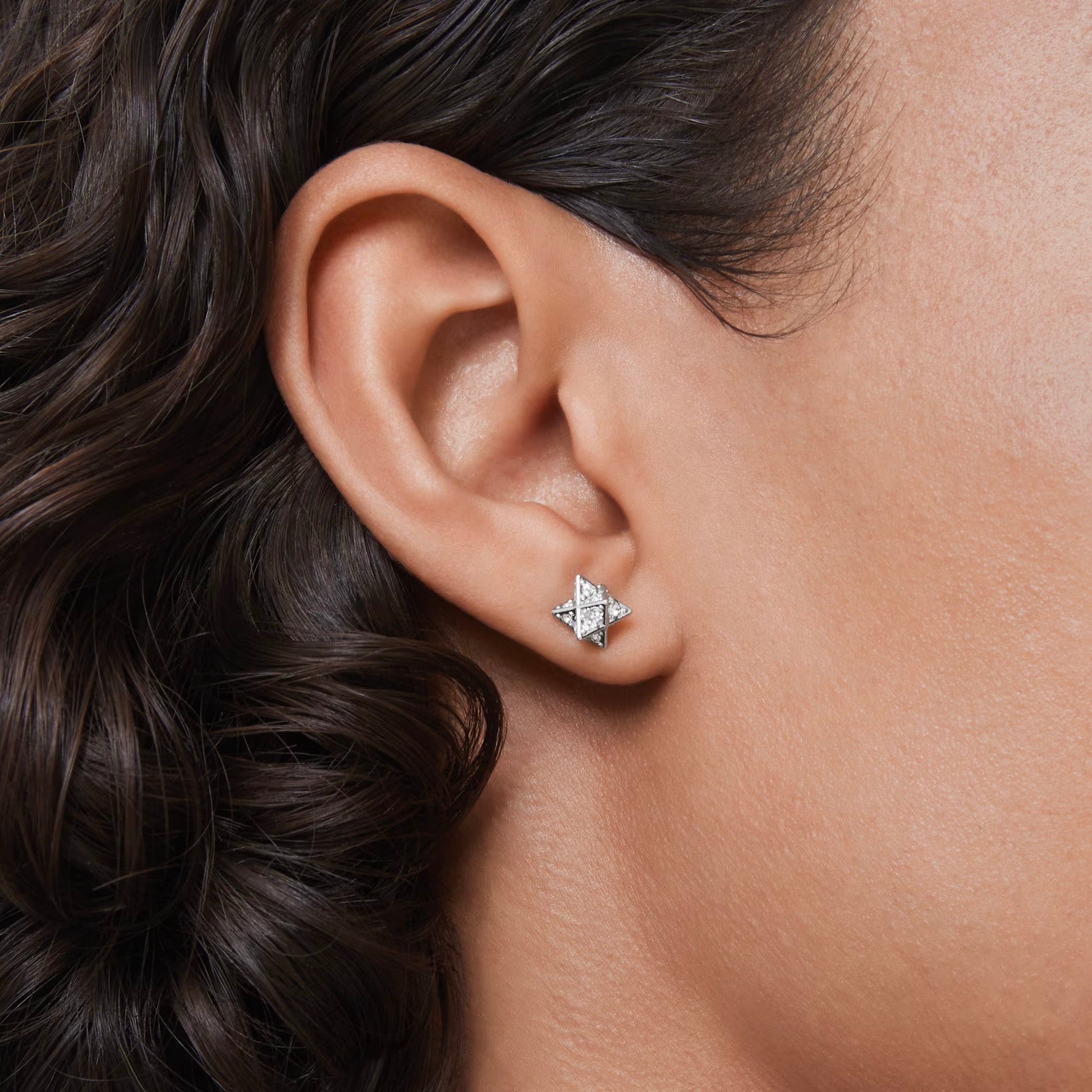 Close-up of a person's ear adorned with a Swarovski Dextera Star Stud Earring, rhodium plated for extra shine. The neutral background enhances the earring and complements the person's curly hair.