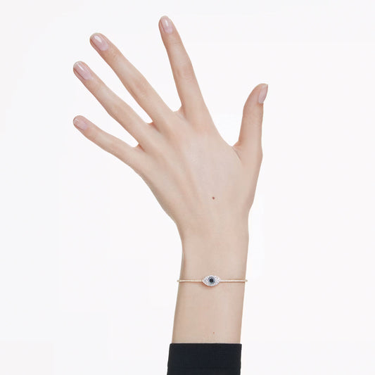 A hand with manicured nails is raised against a white background, showcasing the Swarovski Symbolica Bangle. The delicate gold-tone bracelet from Swarovski features a small, circular charm adorned with a central blue stone. The black sleeve provides an elegant contrast that accentuates the shimmer of the pavé crystals.
