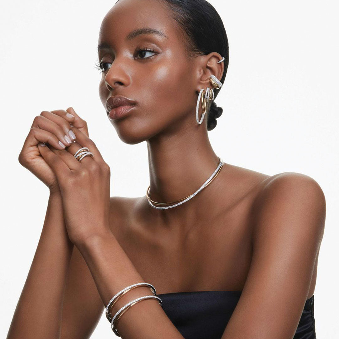 A woman with smooth, glowing skin poses elegantly, adorned in silver jewelry featuring Swarovski Zirconia. She wears the stunning Swarovski Hyperbola Hoop Earrings along with a choker necklace, a ring, and a bracelet. Her hair is pulled back as she graces the lens in her strapless black top against a plain background.
