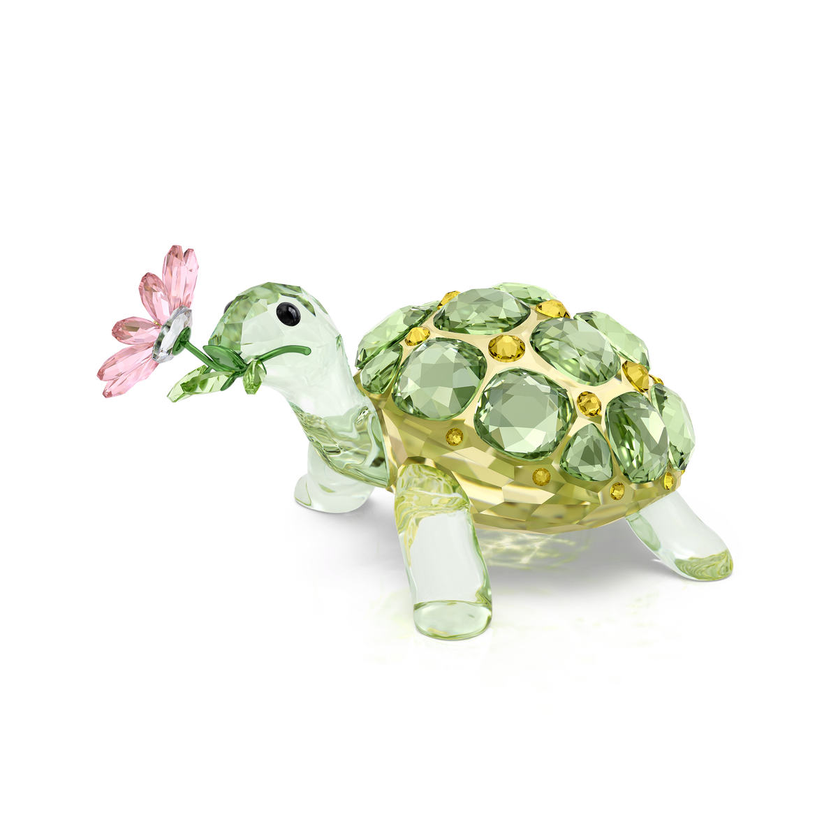 The Swarovski Idyllia Tortoise and Daisy by Swarovski features a small decorative tortoise with a clear glass body and green jeweled shell, holding a pink crystal daisy in its mouth. The sculpture stands on four legs against a plain white background.