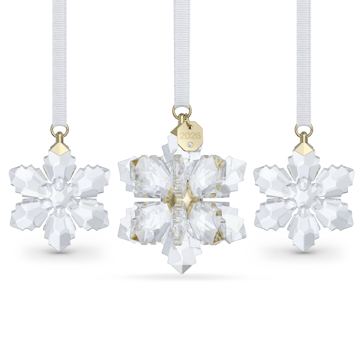 Three Swarovski crystal snowflake ornaments with gold-toned details and white ribbons. The center ornament, from the Swarovski Annual Edition 3D Ornament Set 2026, features a gold tag marked “2026” and is part of the Annual Edition snowflake collection. All three are displayed against a white background.