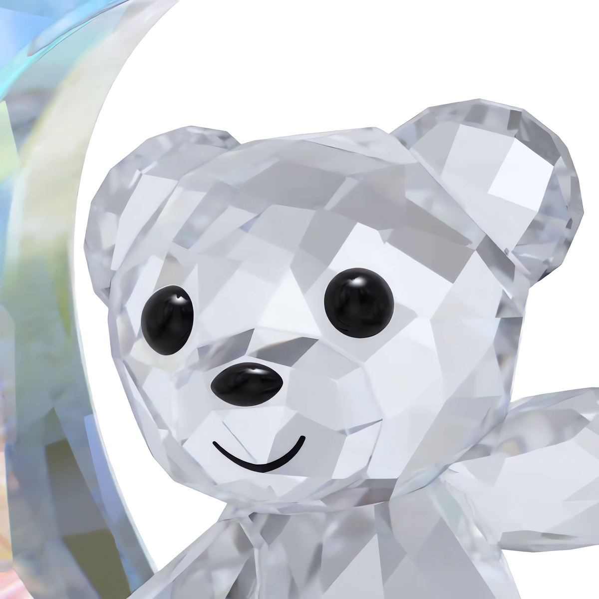 A close-up of the Swarovski Kris Bear Moon Ornament—a faceted crystal figurine with sparkling black eyes and a cheerful smile—set against a plain white background with a touch of rainbow-colored crystal on the left, this charming ornament by Swarovski dazzles playfully in the light.