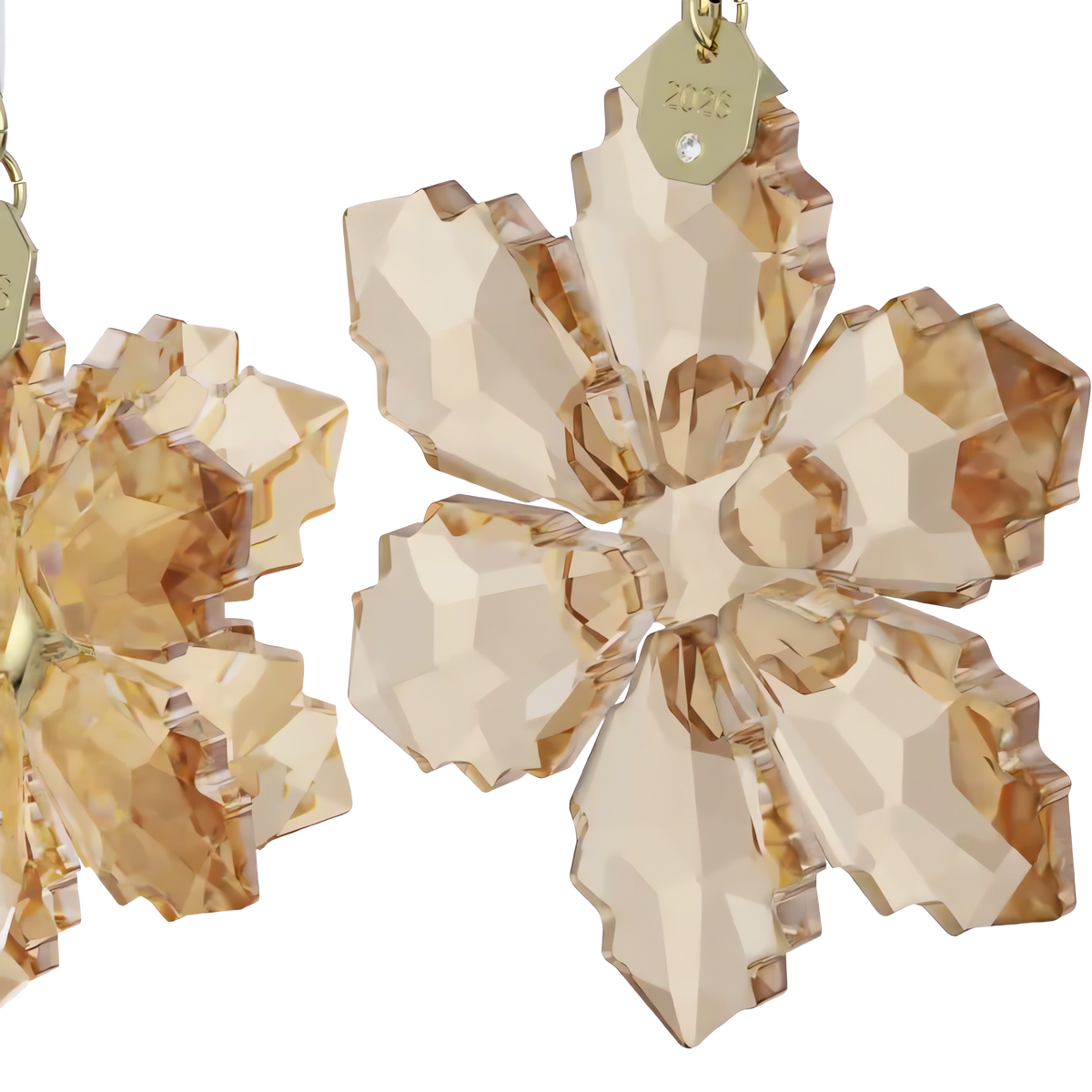 Close-up of two faceted crystal snowflake ornaments in warm amber tones, including a dazzling ornament with gold-colored metal tags—one engraved with “2026.” The Swarovski Annual Edition Festive 3D Ornament Set 2026 is beautifully showcased against a plain white background.