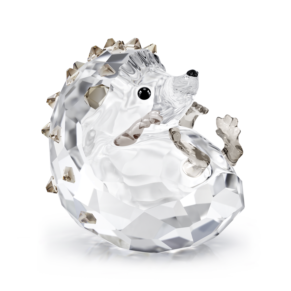 The Swarovski Idyllia Hedgehog by Swarovski is a crystal figurine depicting a curled hedgehog with faceted surfaces, showcasing a clear body paired with light brown transparent spikes and paws. Its shiny black eyes and nose evoke the charm of an autumn forest, all set against a plain white background.