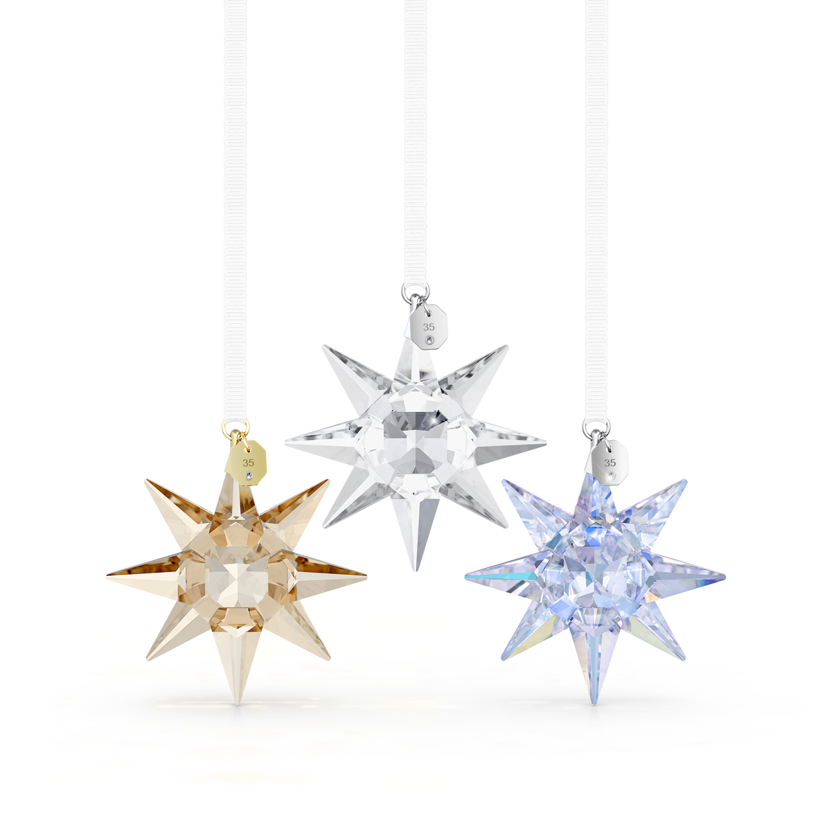 Three sparkling star ornaments from the Swarovski Annual Edition 35th Anniversary Ornament Set 2026—featuring gold, clear, and blue crystals—hang gracefully from white ribbons against a plain white background. Each Swarovski star, inspired by the mesmerizing Aurora Boreale, is crafted with eight shimmering points and adorned with a small metal tag at the top.