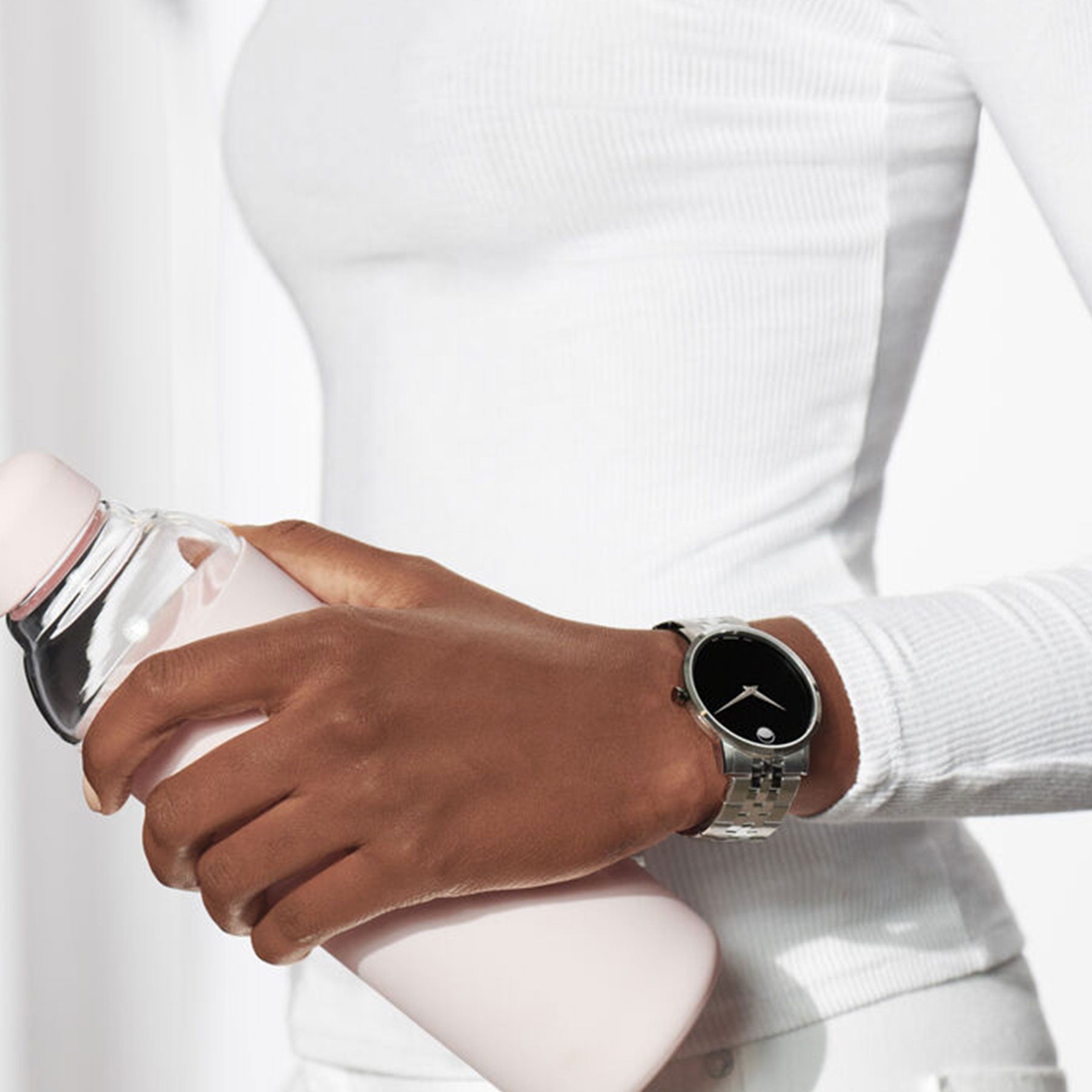 A person wearing a white long-sleeve shirt, holding a water bottle in one hand, showcases their Movado Museum Classic Quartz 40mm Watch with its sleek silver wristwatch visible on their wrist. The bright and minimalistic background highlights the elegance of the stainless steel case design.