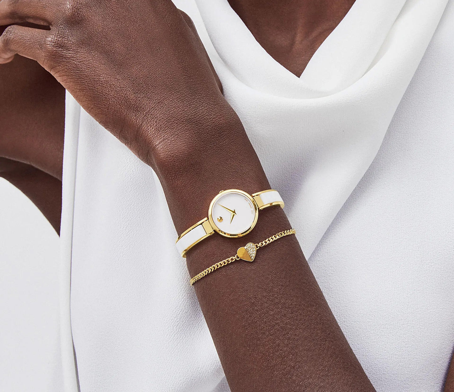 A person wearing a white sleeveless top showcases a yellow gold PVD-finished Movado Moda Quartz 24mm Watch paired with a delicate gold bracelet featuring a heart charm. The watch, with its minimalist Museum dial and white face, perfectly complements the ensemble.