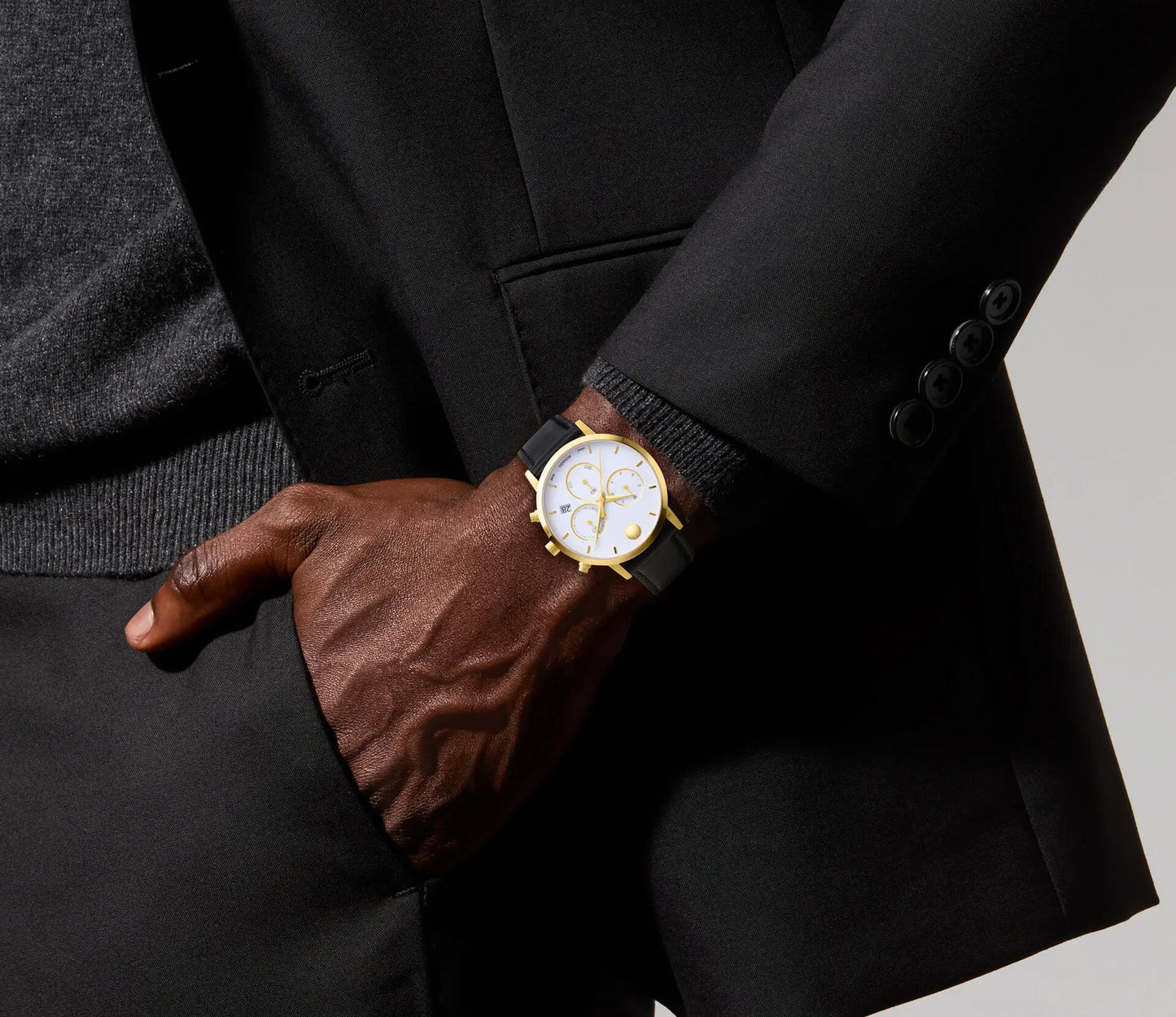 A person in a dark suit with a textured sweater showcases the modern design of the Movado Museum Classic Quartz 42mm Watch. With their hand in the pant pocket, the sleek outfit is complemented by the watch's museum dial in gold and silver tones.