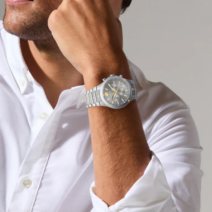 A man in a white shirt showcases the Movado SE Solar 42mm Watch, a sport-elegant timepiece with Swiss craftsmanship. This watch from Movado features a silver case and a gray and yellow dial. He rests his chin on his hand against a neutral background, emphasizing the watch’s perfect combination of precision and style.