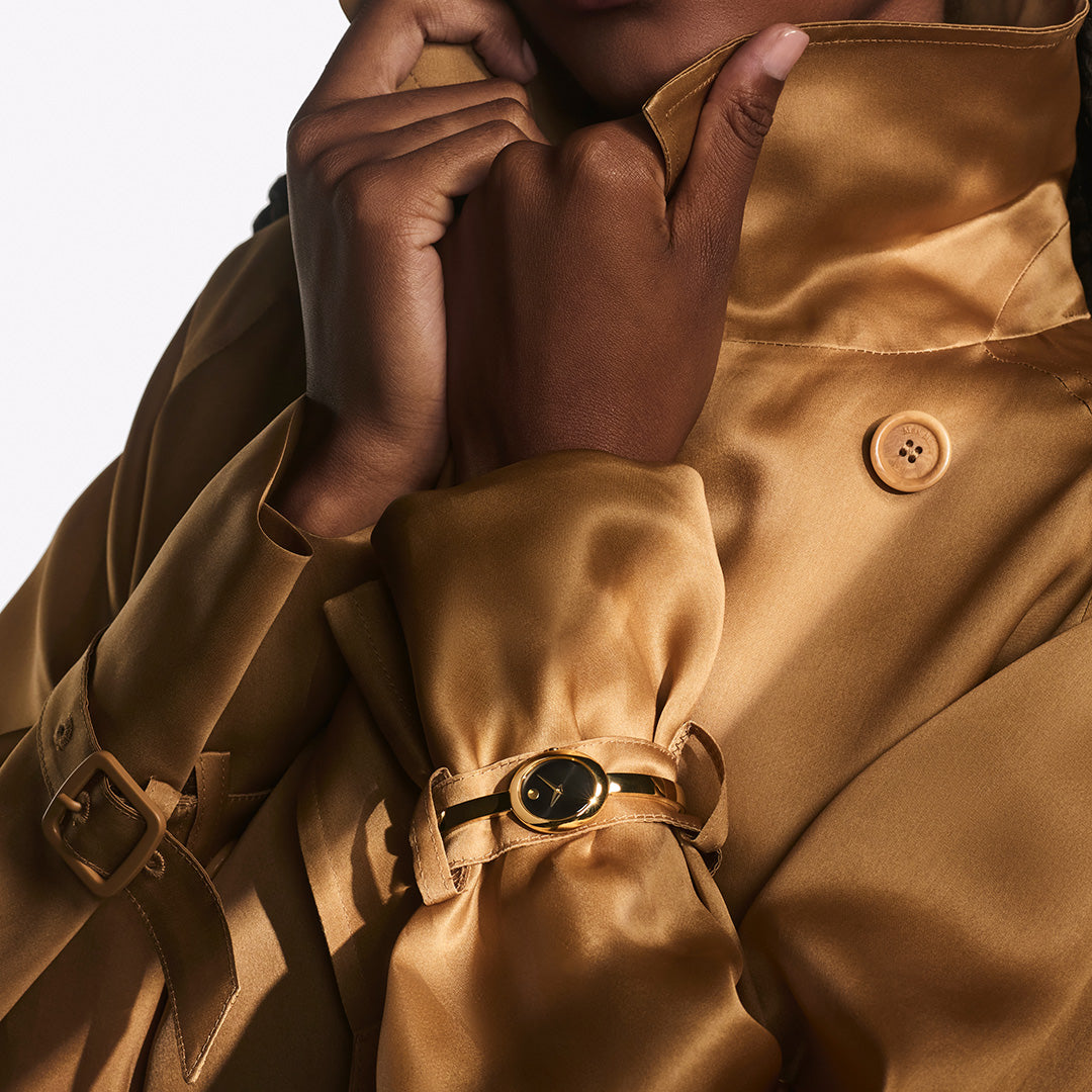 A person wearing a yellow gold PVD Movado Museum Bangle Quartz 21.5mm x 25.5mm Watch with a simple black dial over the sleeve of a tan trench coat.