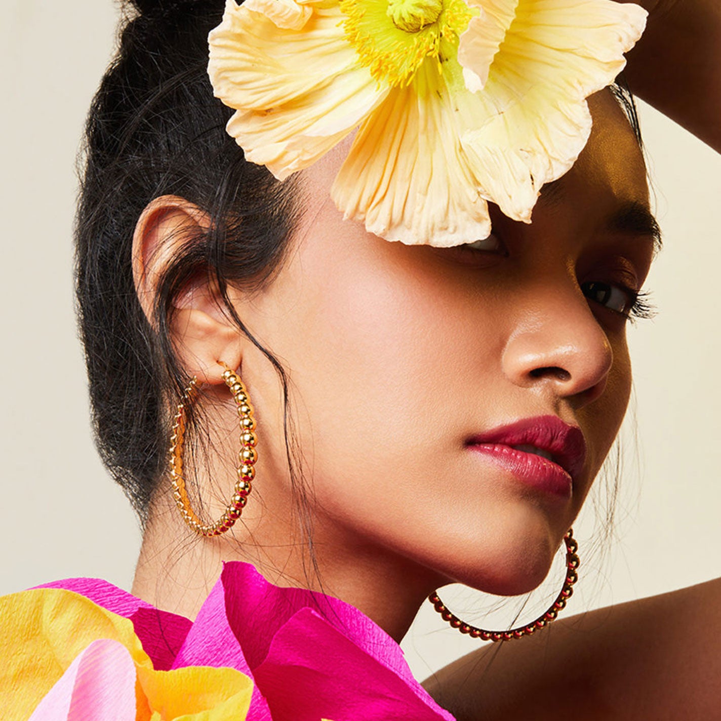 A woman gazes upward, adorned in Roberto Coin Designer 18K Yellow Gold Bead Extra Large Hoop Earrings. Her dark hair is elegantly pulled back, complemented by a large yellow flower. Bright pink petals drape around her neck, harmonizing with her bold pink lipstick.