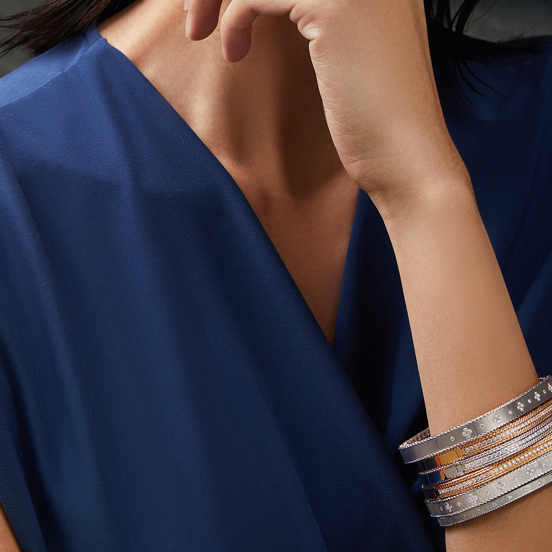 A person in a deep blue V-neck top rests their hand near their chin, displaying stacked metallic bracelets, including the Roberto Coin Princess Satin Fleur De Lis 18K Yellow Gold Diamond Slim Bangle.