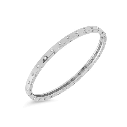 The Roberto Coin Pois Moi 18K White Gold Oval Bangle is a sophisticated bracelet featuring a sleek, reflective finish adorned with uniformly spaced pyramid-shaped studs encircling its surface. Its understated and elegant design is elegantly displayed against a plain white background.