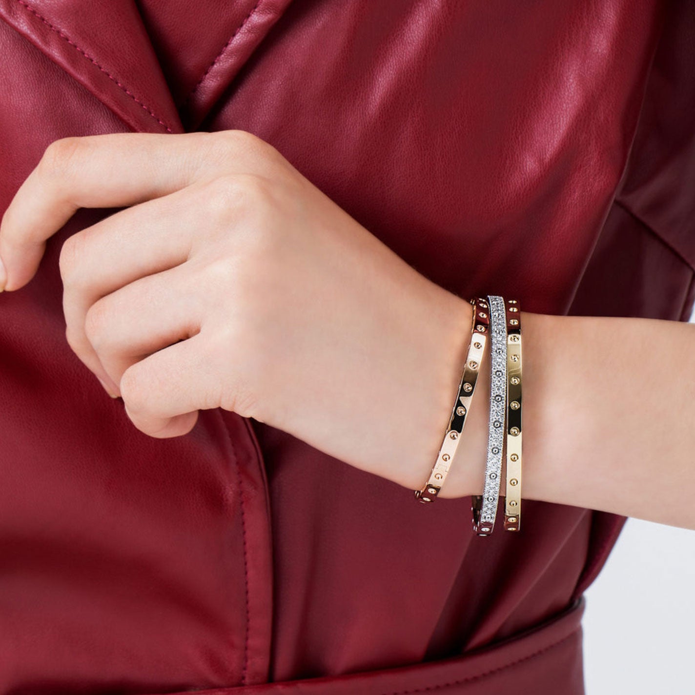 A person dressed in a red leather jacket is displaying their wrist adorned with three elegant bracelets, including the Roberto Coin Pois Moi 18K White Gold Oval Bangle, along with gold, diamond, and burgundy pieces.