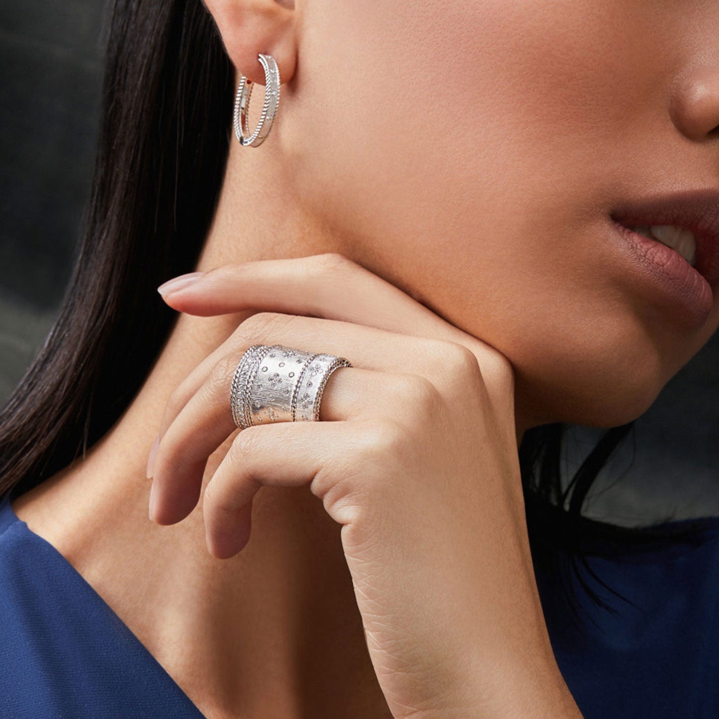 A woman wearing a large, intricately designed 18K white gold ring on her finger and Roberto Coin's Princess Diamond Satin Earrings. Her fingers are gently touching her chin, and she is wearing a dark blue top.