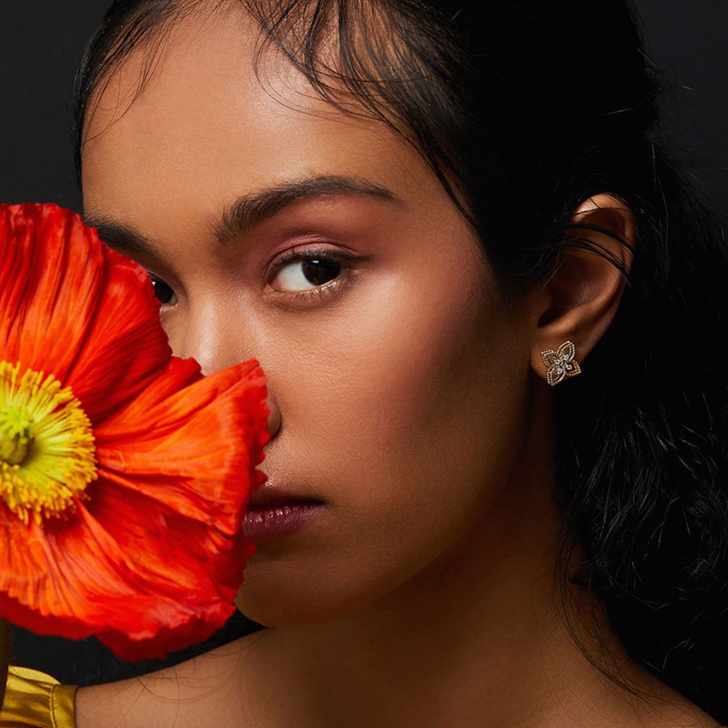 A person with long dark hair holds a large red poppy flower, partially concealing their face, while wearing the dazzling Roberto Coin 18K White Gold Diamond Princess Flower Stud Earrings. They exhibit a serene expression against a black backdrop that enhances the bright colors of the flower and their warm-toned makeup.