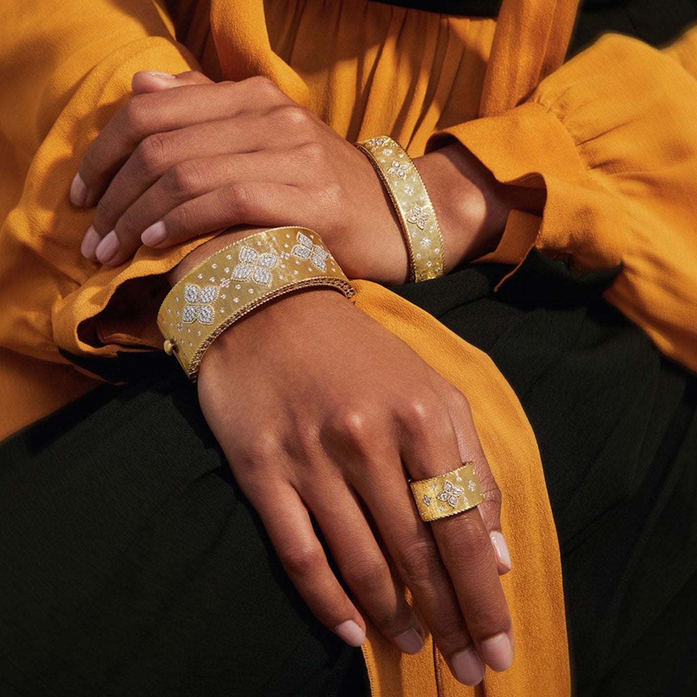 A person wearing a mustard yellow blouse elegantly displays intricate 18k yellow gold jewelry, featuring the Roberto Coin Petite Venetian Princess 18K Yellow Gold Single Satin Flower Diamond Accent Bracelet and a ring adorned with detailed patterns. The person's hands rest on dark fabric, enhancing the sophistication of the diamond-studded accessories.