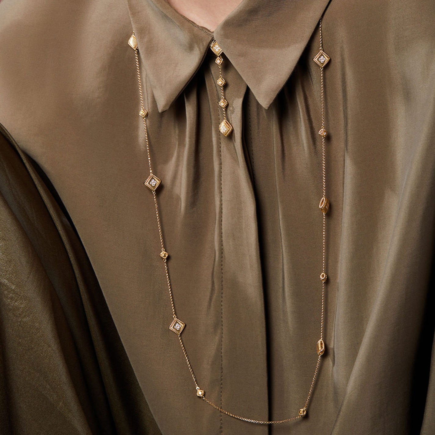 A person is wearing a silky, brown collared blouse complemented by the Roberto Coin Palazzo Ducale 18K Yellow Gold Diamond Station Necklace. The chic chain elegantly lays over the blouse, featuring gold and diamond-shaped pendants that enhance the outfit's sophistication.