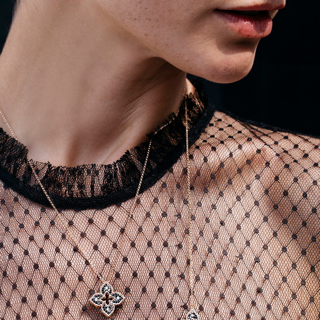 A close-up shows a person in a sheer black polka dot top with a ruffled neckline, wearing two delicate necklaces, including the Roberto Coin Petite Venetian Princess 18K Rose Gold Black and White Diamond Flower Pendant. Their lower face and neck are visible.