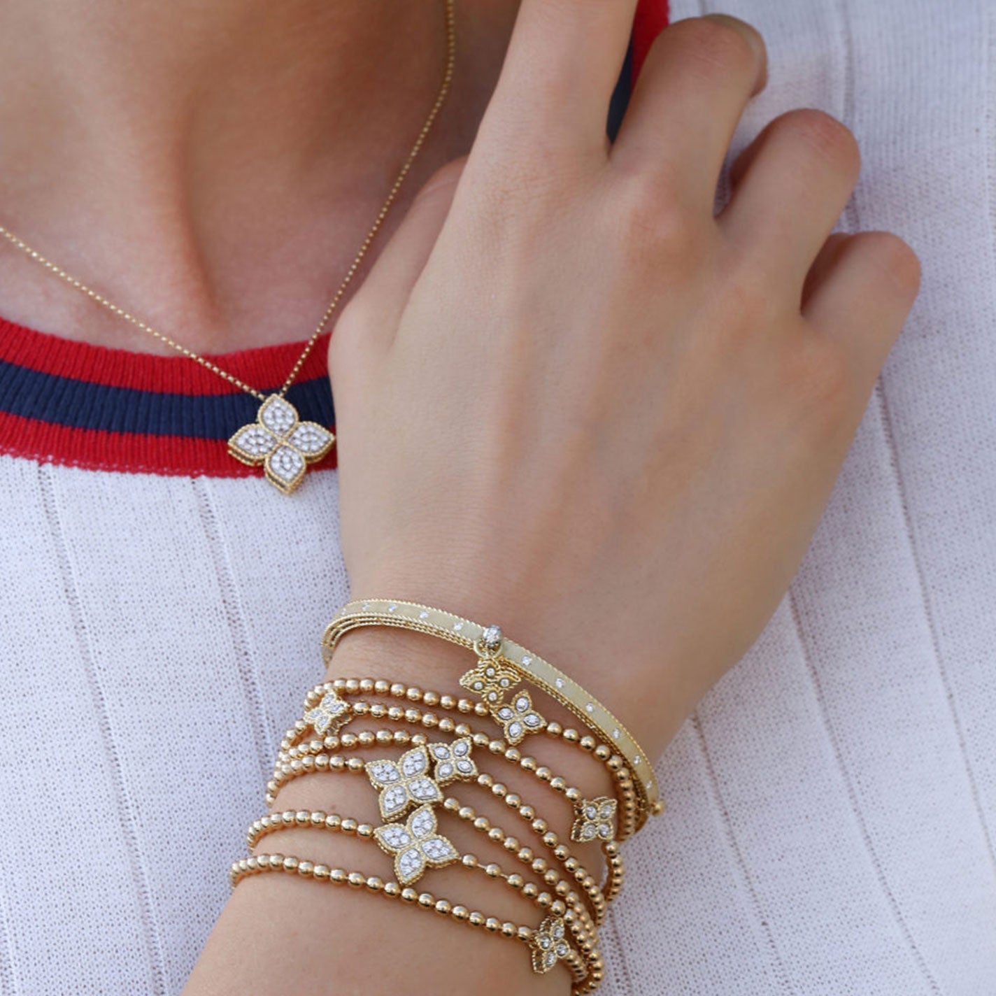 A person in a red and white top is showcasing gold bracelets and a necklace adorned with floral-shaped, diamond-studded charms from Roberto Coin. The raised hand highlights the Roberto Coin Medium Princess Flower 18K Yellow and White Gold Diamond Stretch Bracelet against the fabric.