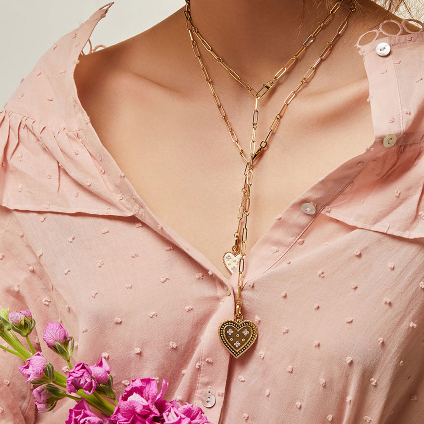 A person in a pink textured blouse is accessorized with a layered gold chain necklace, showcasing heart-shaped pendants and a radiant element from the Roberto Coin Venetian Princess 18K Yellow Gold Large Diamond Heart Medallion Necklace Pendant. In their hand, they hold a bouquet of pink flowers at the bottom left of the image.