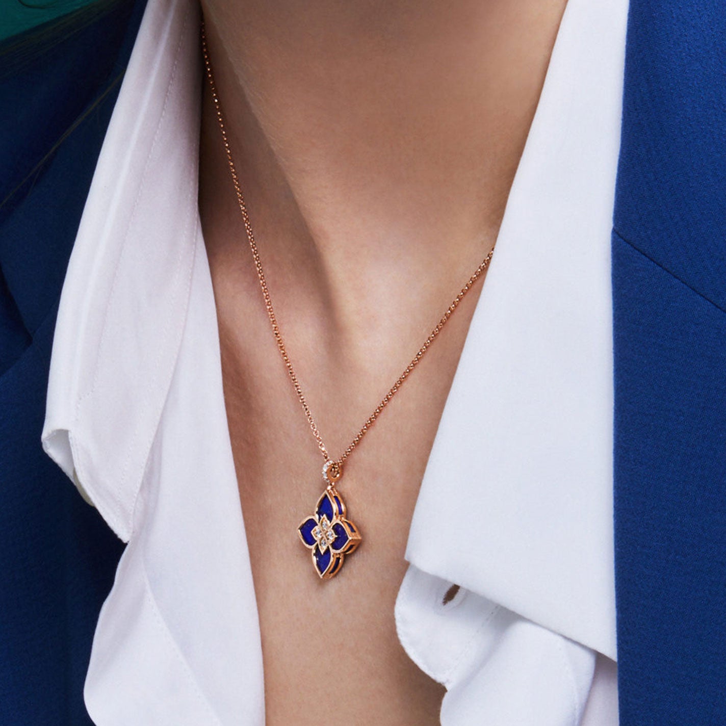 A woman wearing a blue blazer and an open-collar white shirt showcases a Roberto Coin necklace. The Venetian Princess Pendant, crafted from 18 Karat Rose Gold, features a beautiful four-petal flower adorned with blue lapis and diamond accents.