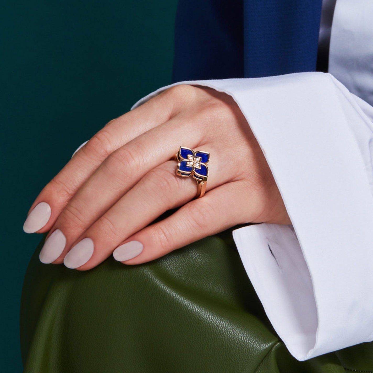 The hand with manicured nails is adorned with a Roberto Coin Venetian Princess 18K Rose Gold Small Lapis and Diamond Ring. The person is wearing a white shirt, and the sleeve of their blue blazer is partially visible as they hold a green leather item.