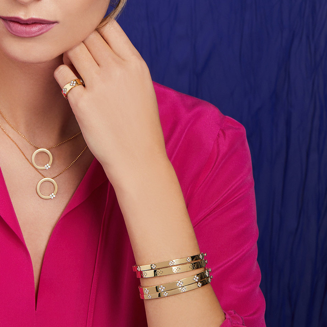 A person in a pink blouse showcases jewelry, including a gold necklace with pendants, a ring, and the Roberto Coin Love In Verona 18K White Gold Medium Width Bangles with diamond accents and safety clasps, set against a blue background.