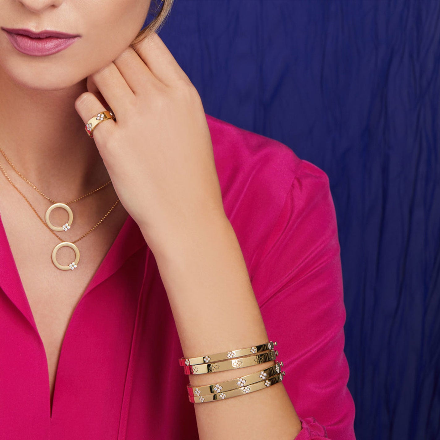 A woman in a vibrant pink blouse elegantly displays her chic accessories, featuring Roberto Coin necklaces with circular pendants. She adorns herself with a ring and several Roberto Coin Love In Verona 18K Rose Gold Medium Width Bangles. The deep blue background highlights her fashionable outfit and jewelry inspired by the Love In Verona collection.