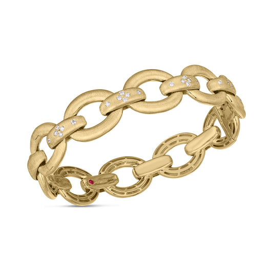 The Roberto Coin Duchessa 18K Yellow Gold with Diamond Accent Satin Oval Link Hinged Bangle features oval links with a brushed finish, accented by diamond clusters on some links, while the inner side is embellished with a smaller link featuring a tiny red gemstone.