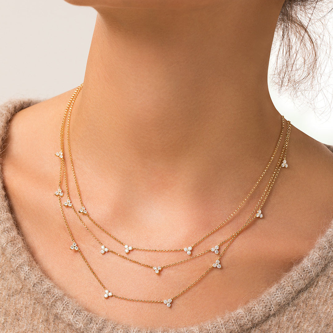 A close-up of a woman’s neck wearing a beige knit sweater and the Roberto Coin Diamonds by the Inch 18K Yellow Gold Diamond Station Flower Necklace, featuring diamond clusters on a delicate 17" chain. The background is softly lit.