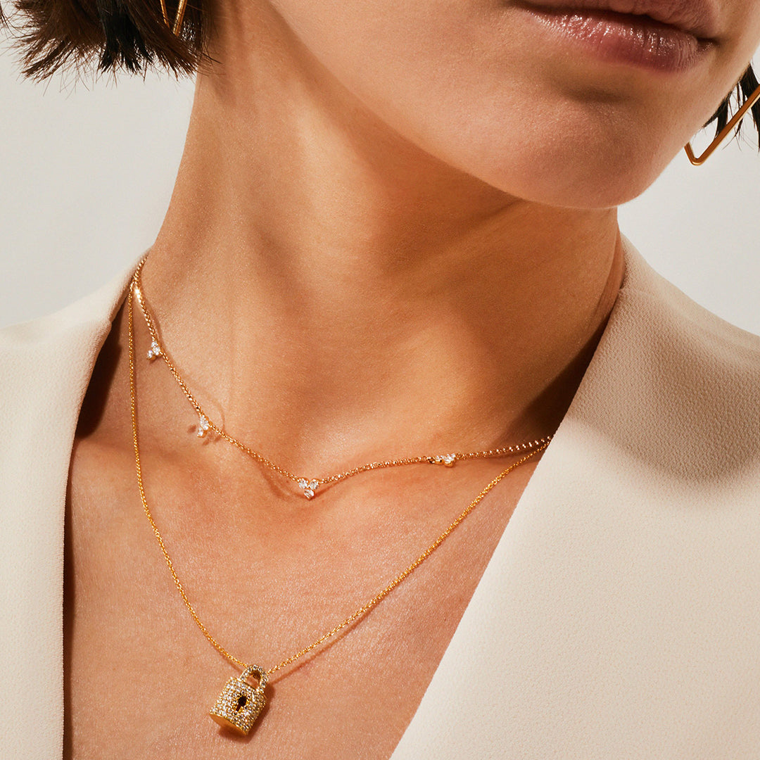 A close-up shows a woman in a cream blazer wearing two gold necklaces: the Roberto Coin Diamonds by the Inch 18K Yellow Gold Diamond Station Flower Necklace and a 17" lock pendant. Her short hair frames parted lips.