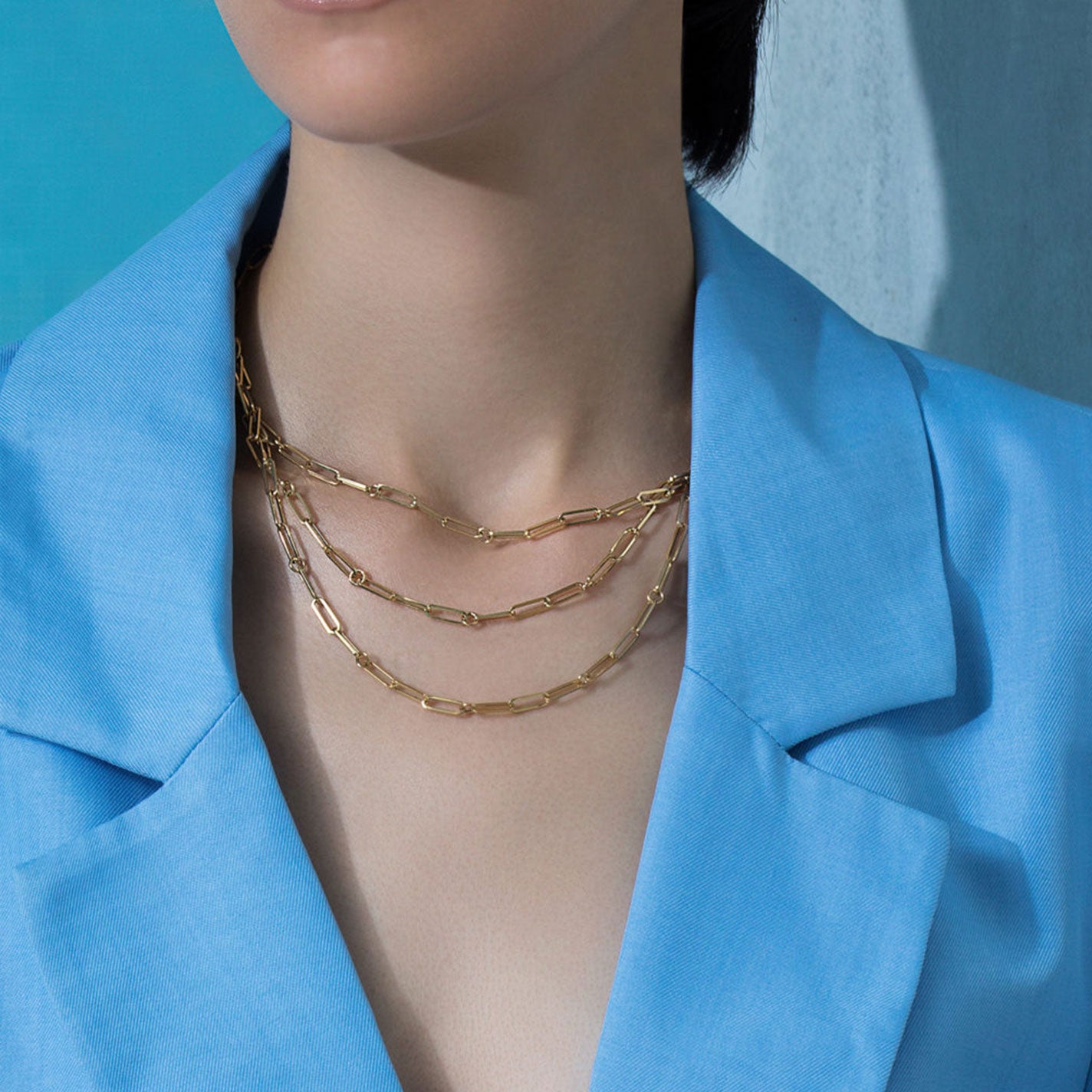 A person in a blue blazer is adorned with layered chains, featuring the exquisite Roberto Coin Designer 18K Yellow Gold Triple Strand Paperclip Chain Diamond Necklace. The backdrop of blue and gray adds an elegant contrast.