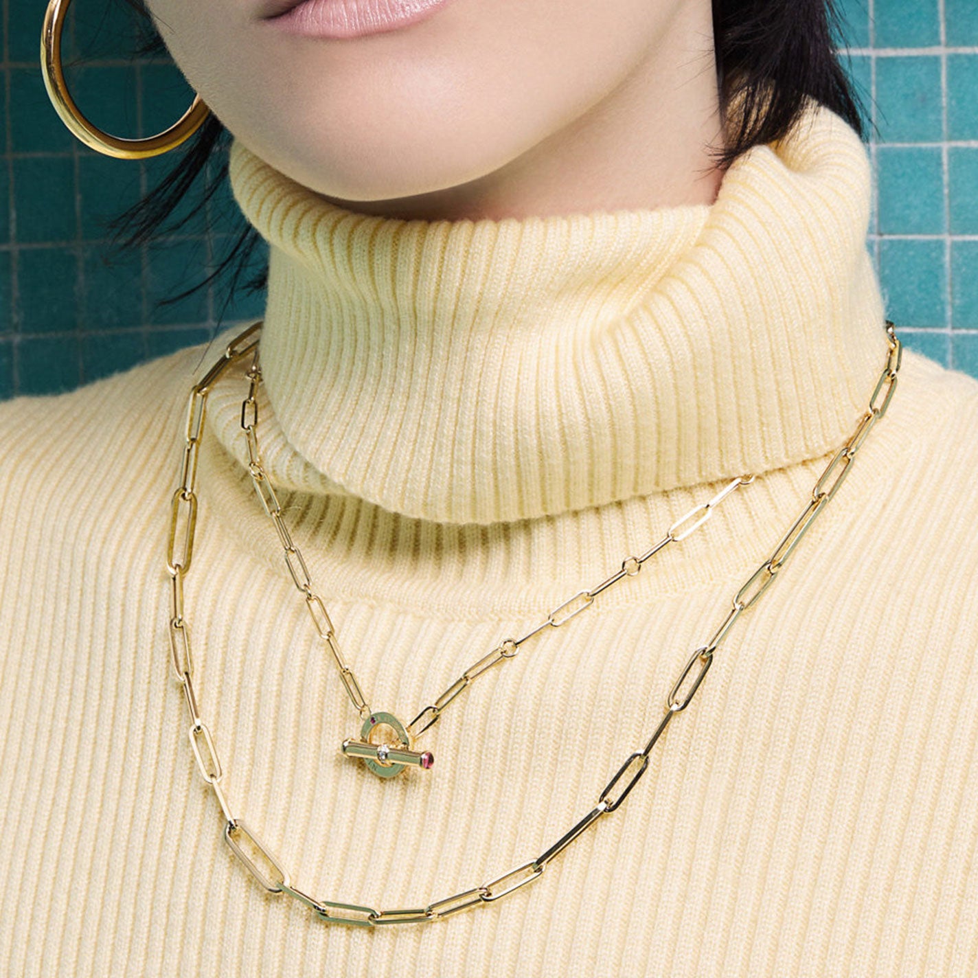 A person wearing a ribbed, cream-colored turtleneck sweater is adorned with two gold chain necklaces. One necklace is the Roberto Coin Designer 18K Yellow Gold Paperclip Chain Necklace, featuring a distinctive diamond toggle closure with a small red gem. The background is a teal, tiled wall.