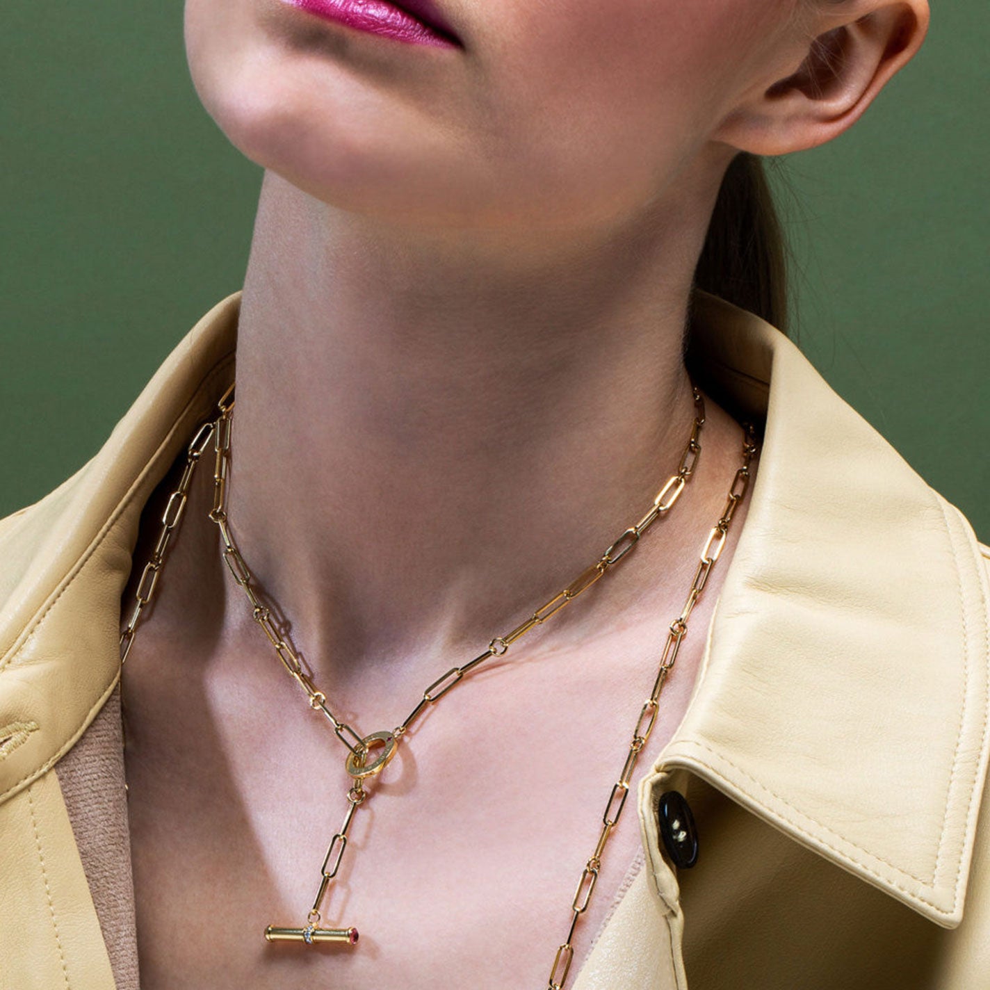 Against a green background, a person showcases the elegance of their attire, featuring a light beige trench coat complemented by two layered gold chain necklaces. One is the Roberto Coin Designer 18K Yellow Gold Paperclip Chain Diamond Toggle Closure Necklace, highlighting exquisite craftsmanship and style.