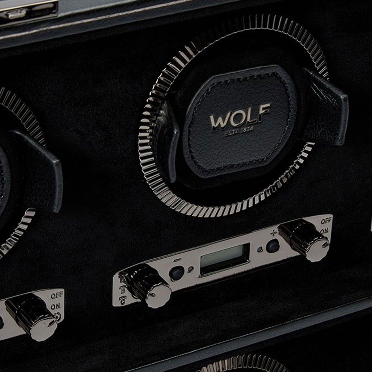 A close-up of the WOLF 1834 British Racing 8PC Watch Winder reveals a timeless design, with "WOLF EST. 1834" elegantly inscribed on a prominent component. The watch winder features round holders and metallic control knobs, complemented by a digital display for settings, all embodying the essence of British Racing aesthetics. The interior is luxuriously lined in black material.