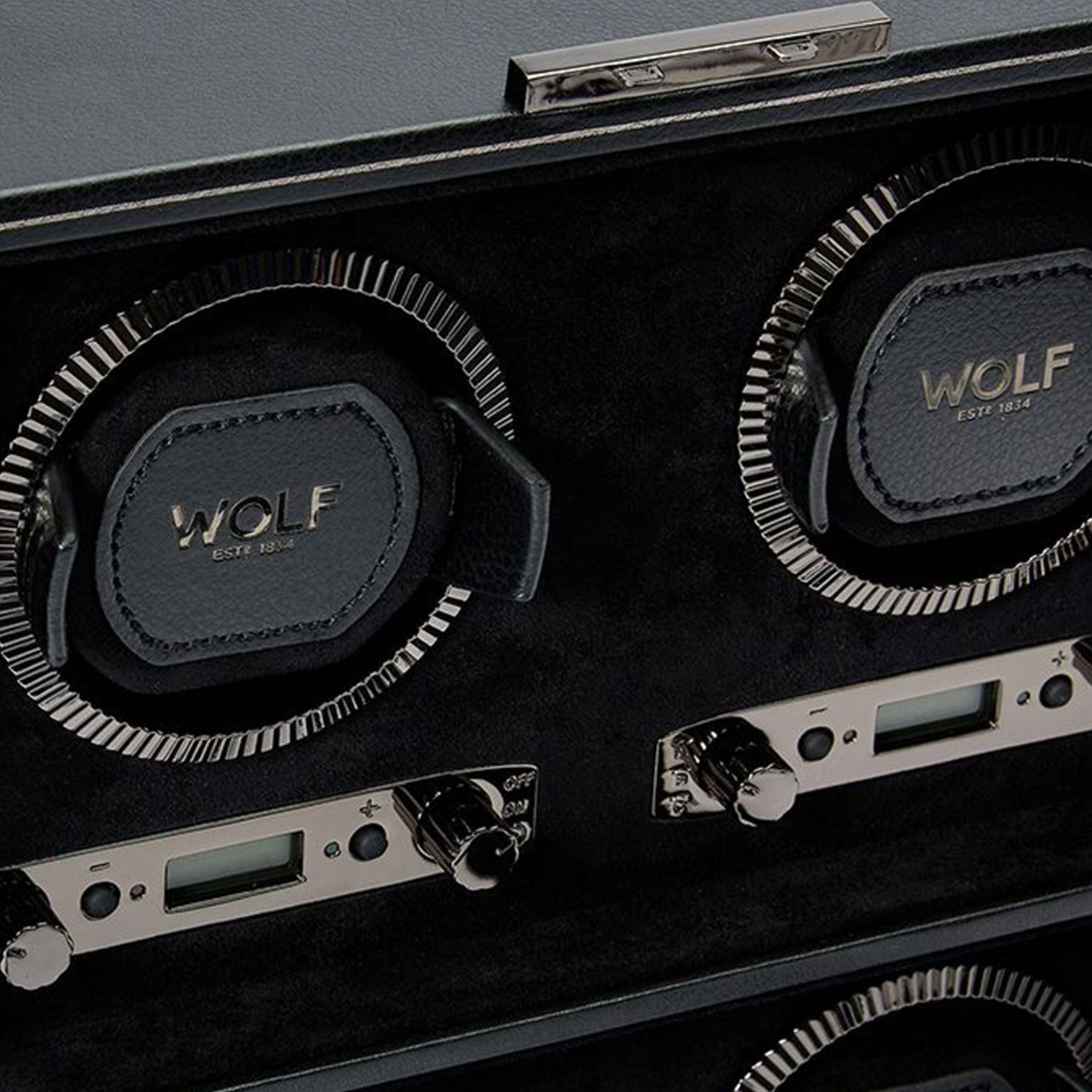 A close-up of the WOLF 1834 British Racing 4PC Watch Winder showcases "Est. 1834" embossed on its black leather interior. This device exudes timeless elegance with silver dials and digital displays, embodying the sophistication of British Racing.