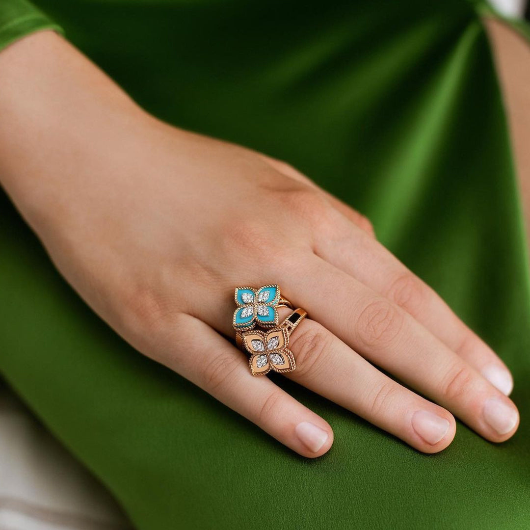A hand rests on a green fabric, featuring two floral rings. The top is a Roberto Coin Venetian Princess 18K Rose Gold Diamond and Turquoise Flower Ring, while the bottom showcases a beige and white flower design in 18 Karat Rose Gold.