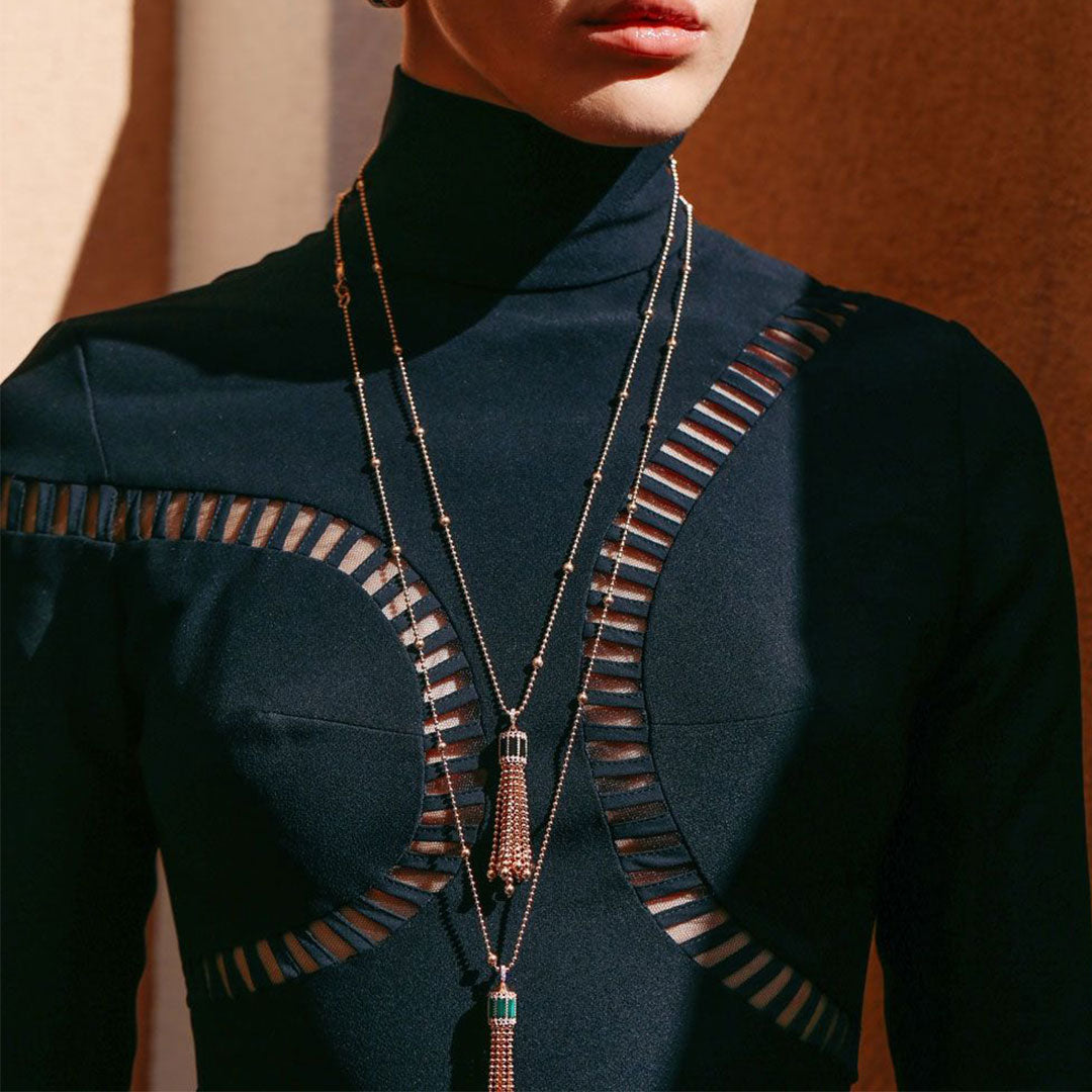 A person wearing a high-neck black top with transparent panel designs is adorned with layered necklaces featuring tassels, including the Roberto Coin Art Deco 18K Rose Gold Malachite with Diamond Necklace Pendant. The sunlight casts interesting shadows on the clothing, highlighting the textures and details.