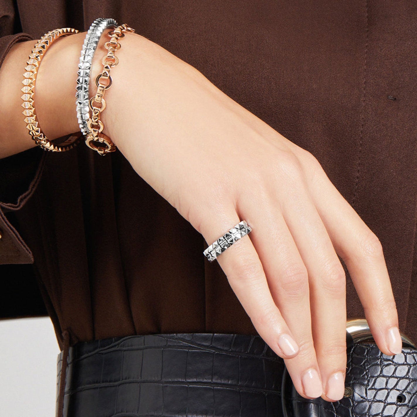 A person wearing a brown blouse and croc-embossed belt showcases a hand adorned with a silver ring and three bracelets: a gold chain, a silver linked design, and the Roberto Coin Obelisco 18K White Gold Hinged Bangle featuring mixed metal heart accents.